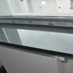 Szary, błyszczący parapet z konglomeratu zamontowany w oknie PCV z widocznym grzejnikiem pod spodem.