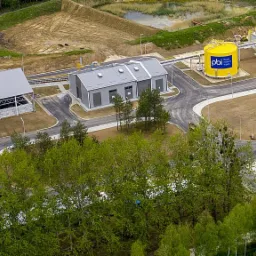 Kompleks budynków przemysłowych z żółtym zbiornikiem gazu widziany z lotu ptaka, otoczony zielenią i utwardzonym terenem, z widoczną drogą dojazdową i zbiornikiem retencyjnym.