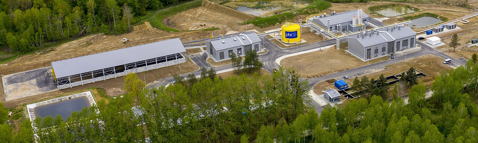 Kompleks budynków przemysłowych z żółtym zbiornikiem gazu widziany z lotu ptaka, otoczony zielenią i utwardzonym terenem, z widoczną drogą dojazdową i zbiornikiem retencyjnym.