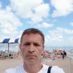 Mężczyzna robiący selfie na zatłoczonej, piaszczystej plaży z widokiem na morze i błękitne niebo z chmurami. W tle widoczne białe parasole i leżaki.