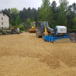Żółty spychacz Mecalac AX 850 z niebieskim osprzętem profilującym teren, w tle drugi spychacz i budynek mieszkalny, teren wyrównany drobnym żwirem, pochmurne niebo.