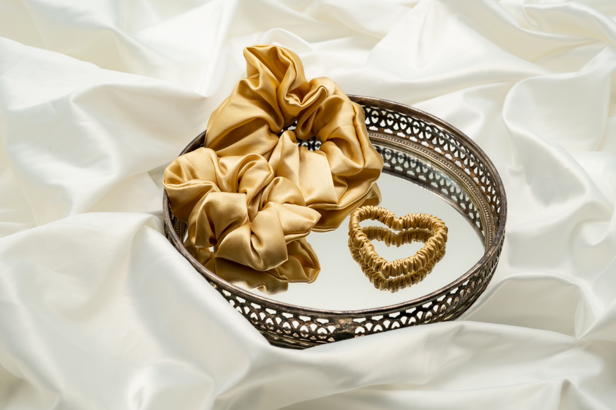 Dwie złote gumki do włosów, jedna w formie serca, leżą na srebrnej, zdobionej tacy z lustrzanym dnem, na tle białej, jedwabnej tkaniny.