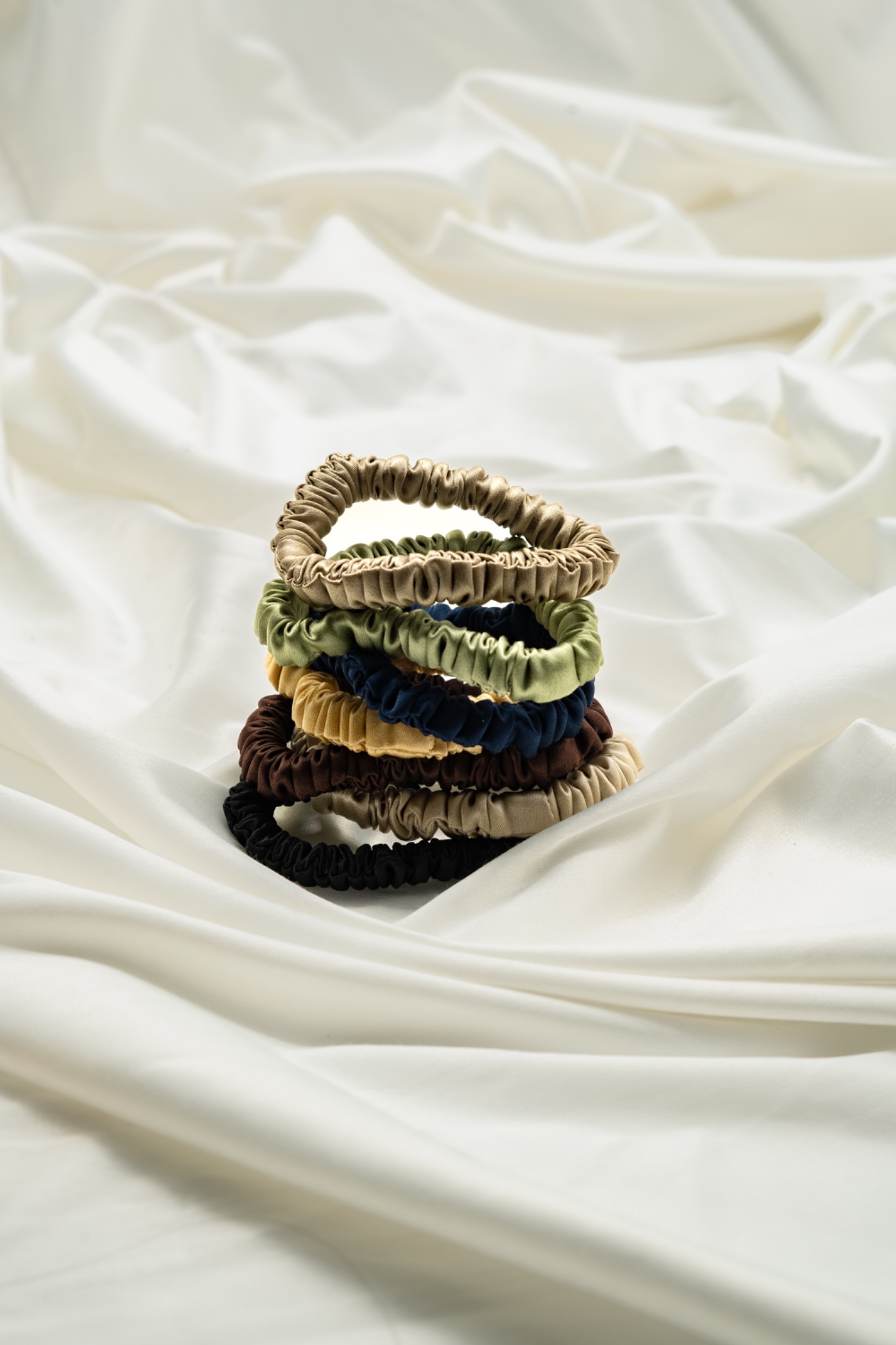 Wieża z jedwabnych gumek do włosów w odcieniach złota, zieleni, granatu, brązu i czerni na tle kremowej, satynowej tkaniny z delikatnymi fałdami.