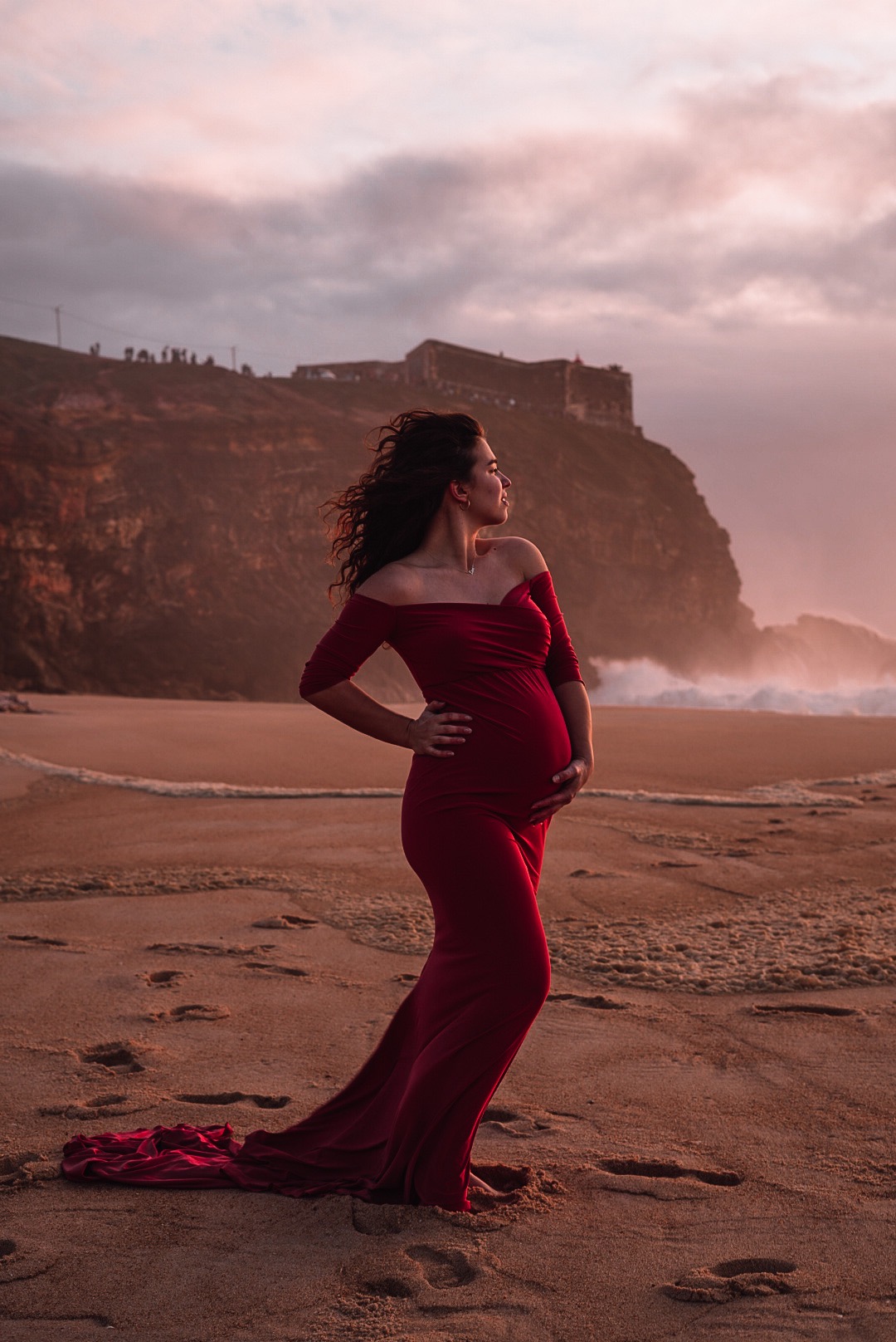 Kobieta w czerwonej sukni ciążowej na plaży w Lubinie, z ręką na brzuchu i wiatrem we włosach, na tle klifu i zachodzącego słońca.