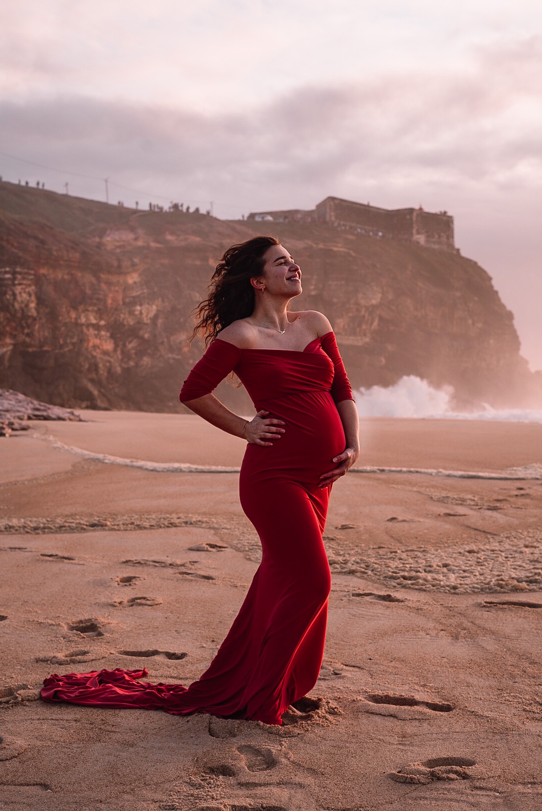 Uśmiechnięta kobieta w czerwonej sukni ciążowej pozuje na plaży z klifem i falami w tle, trzymając ręce na brzuchu.