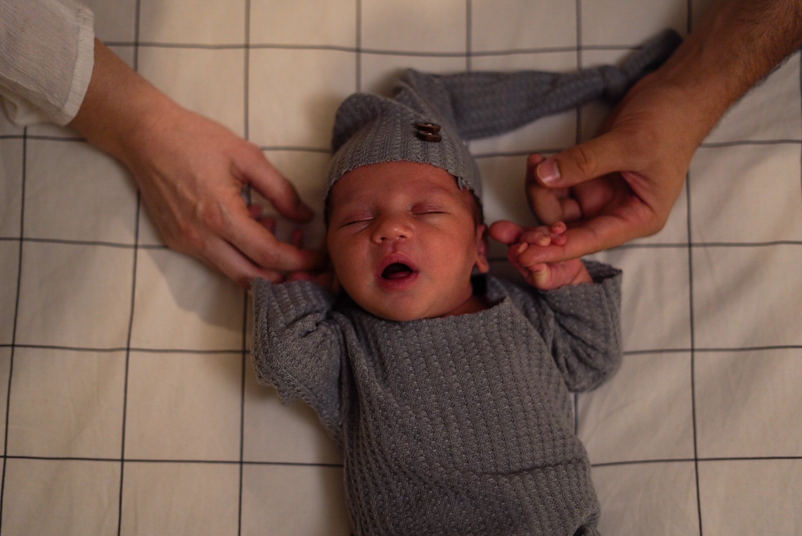 Noworodek w szarym ubranku i czapeczce z guzikiem, leżący na białym tle w kratę, trzymany za rączkę przez rodziców.