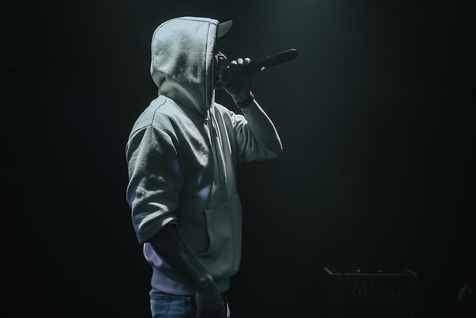 Mężczyzna w bluzie z kapturem i czapce z daszkiem, trzymający mikrofon na scenie w ciemnym otoczeniu.