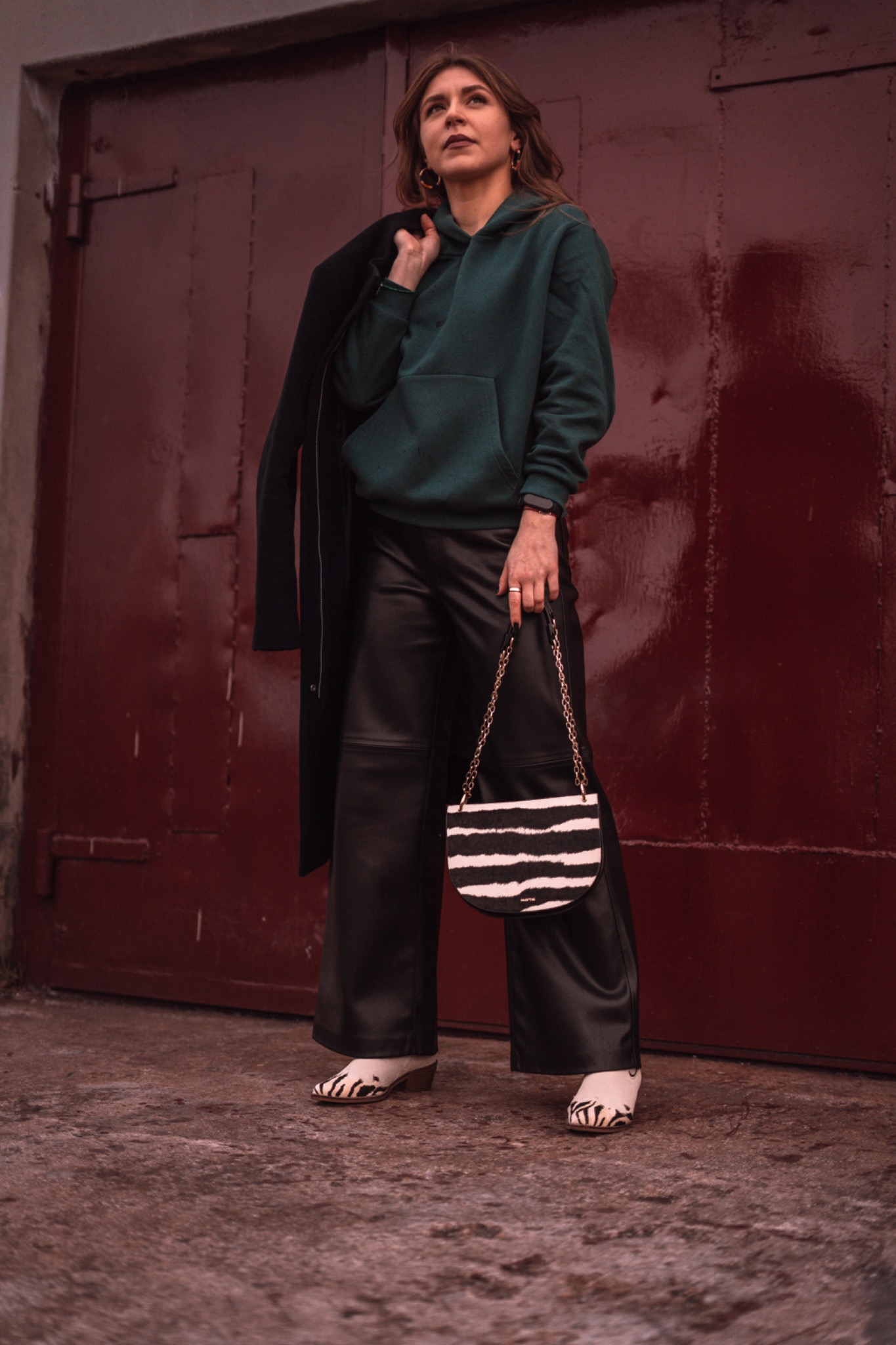Kobieta w zielonej bluzie z kapturem, skórzanych spodniach i butach w zebrę, trzymająca torebkę w paski na tle czerwonych drzwi garażowych.