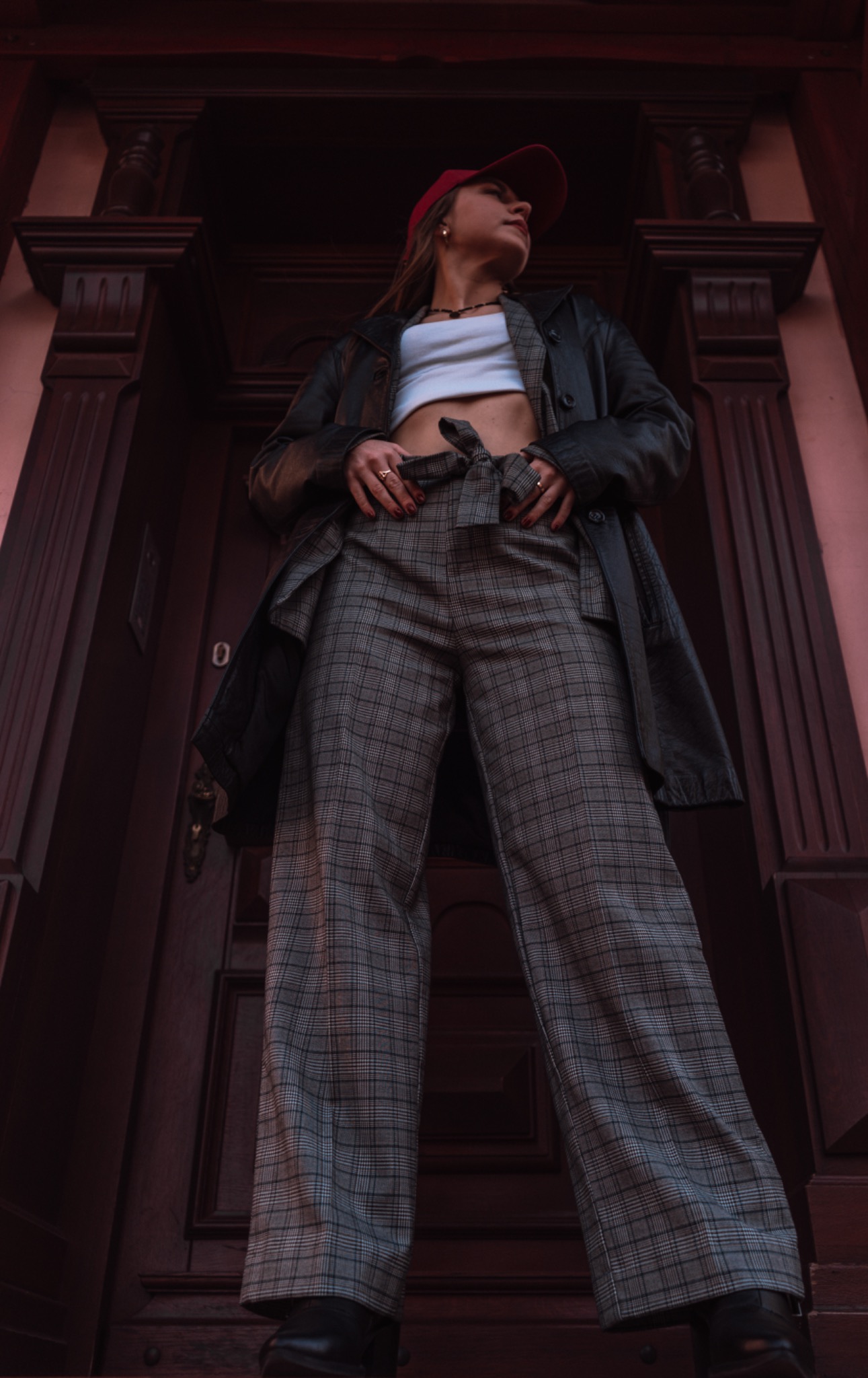 Kobieta w czerwonej czapce, białym topie i spodniach w kratę pozuje przed ciemnymi, drewnianymi drzwiami, patrząc w górę z perspektywy żabiej.