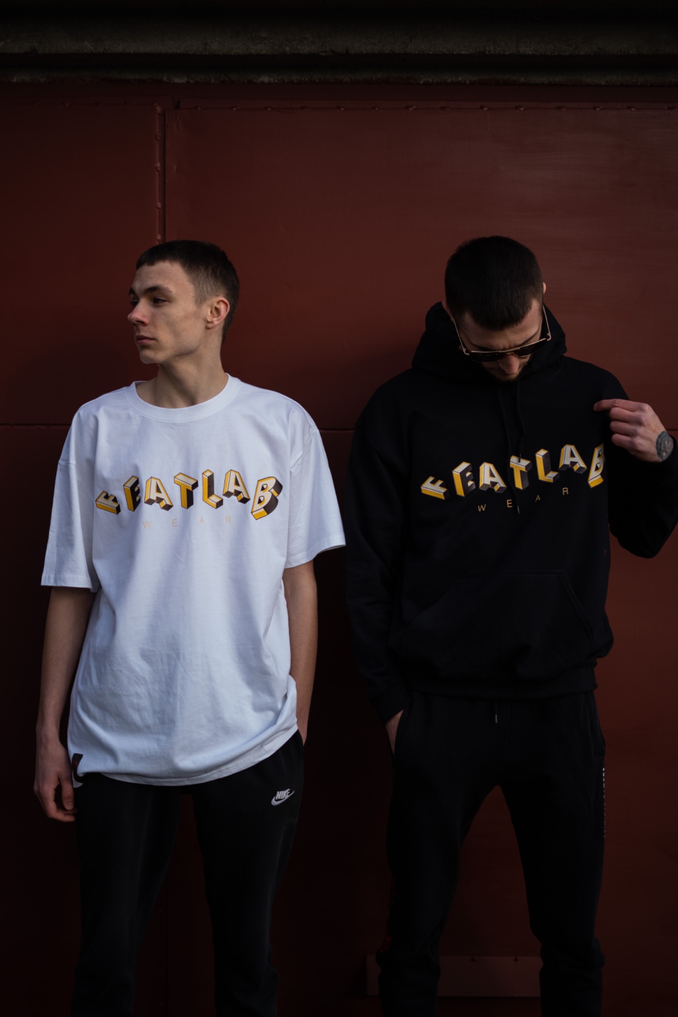 Dwóch mężczyzn pozuje na tle ciemnej ściany w ubraniach z logo 'FATLAB WEAR': jeden w białej koszulce, drugi w czarnej bluzie z kapturem.