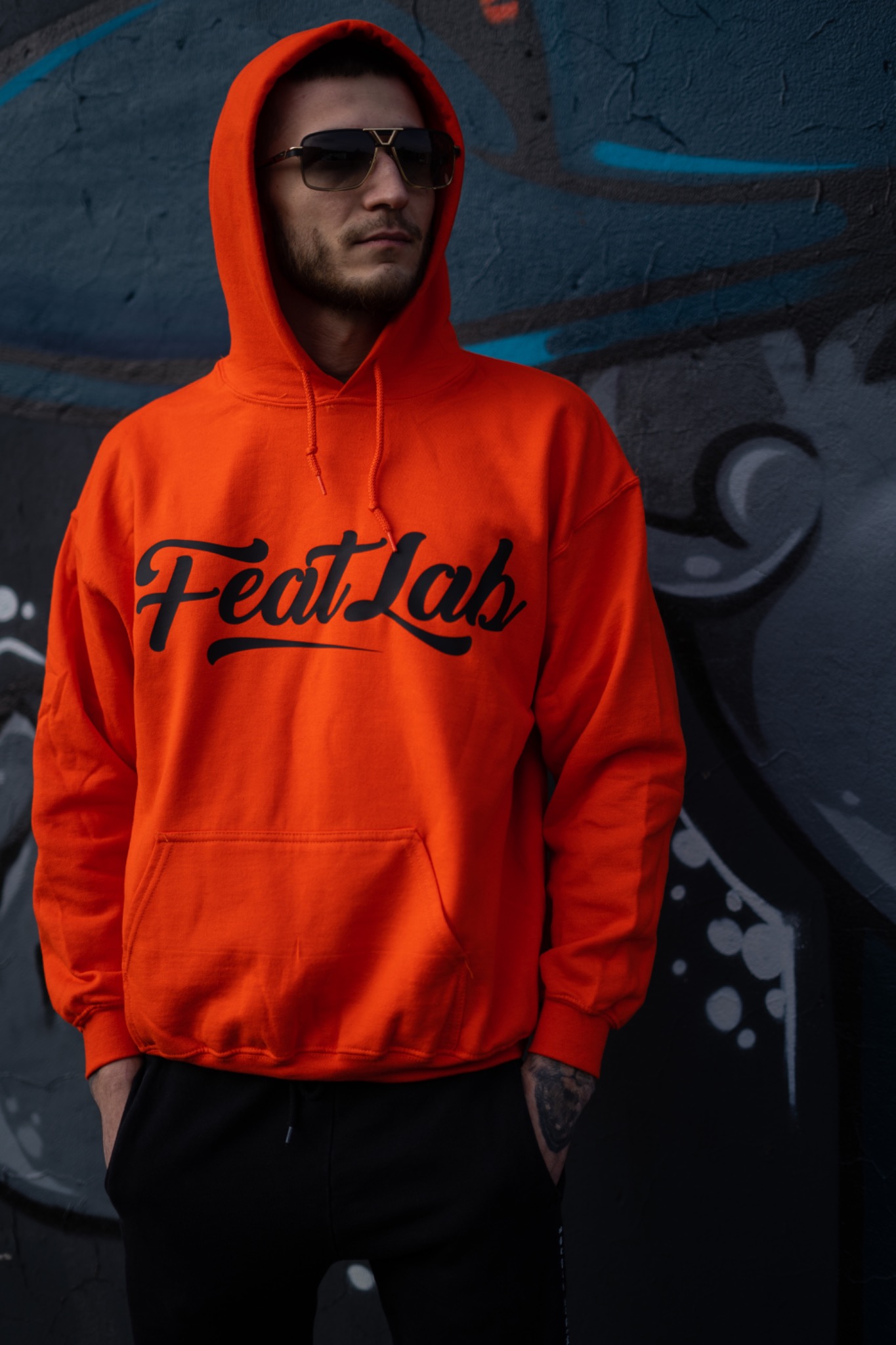 Mężczyzna w pomarańczowej bluzie z kapturem 'Feat Lab' i okularach przeciwsłonecznych, pozujący na tle ściany z graffiti.