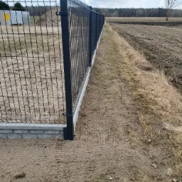 Nowoczesne ogrodzenie panelowe z podmurówką z bloczków betonowych, widok z perspektywy ziemi, w tle pole i drzewa.