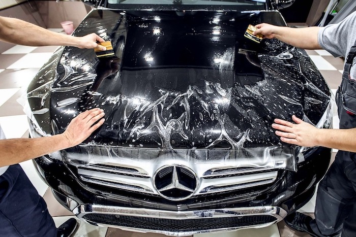 Nakładanie folii ochronnej na maskę czarnego Mercedesa; widoczne dłonie pracowników używających rakli do wygładzania folii.
