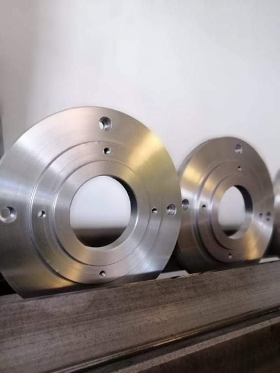 Metalowe, okrągłe elementy z otworami, prawdopodobnie kołnierze, leżące na metalowej półce, po obróbce.