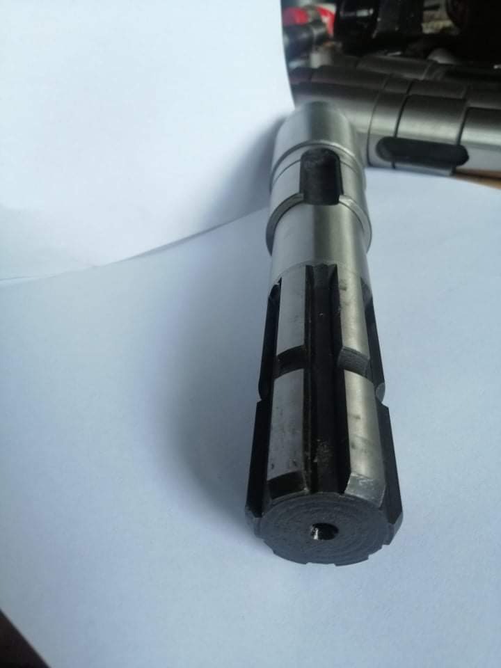 Metalowy wał z rowkami i otworem, leżący na białym tle, widoczne ślady obróbki.