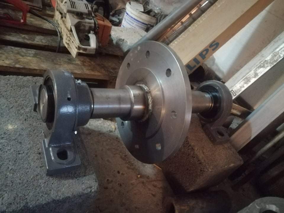 Metalowy wał z kołnierzem i łożyskami zamontowany na betonowych podstawach, w tle piła łańcuchowa i inne przedmioty.