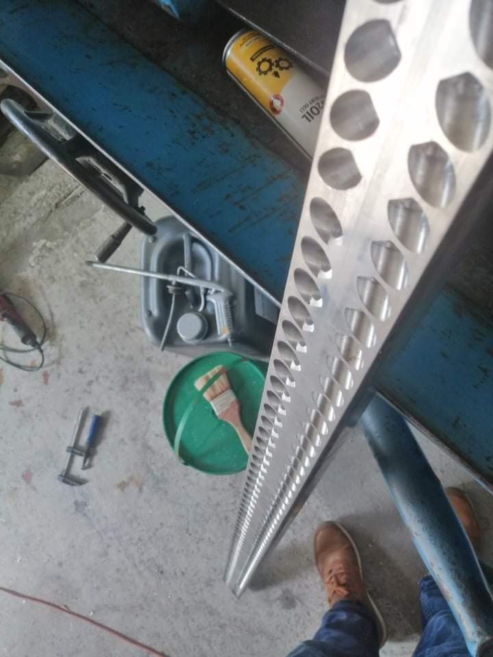 Długa, aluminiowa listwa z rzędem okrągłych otworów, leżąca na niebieskim stole warsztatowym obok puszki oleju, zielonego wiadra z pędzlem i stopy w brązowym bucie.