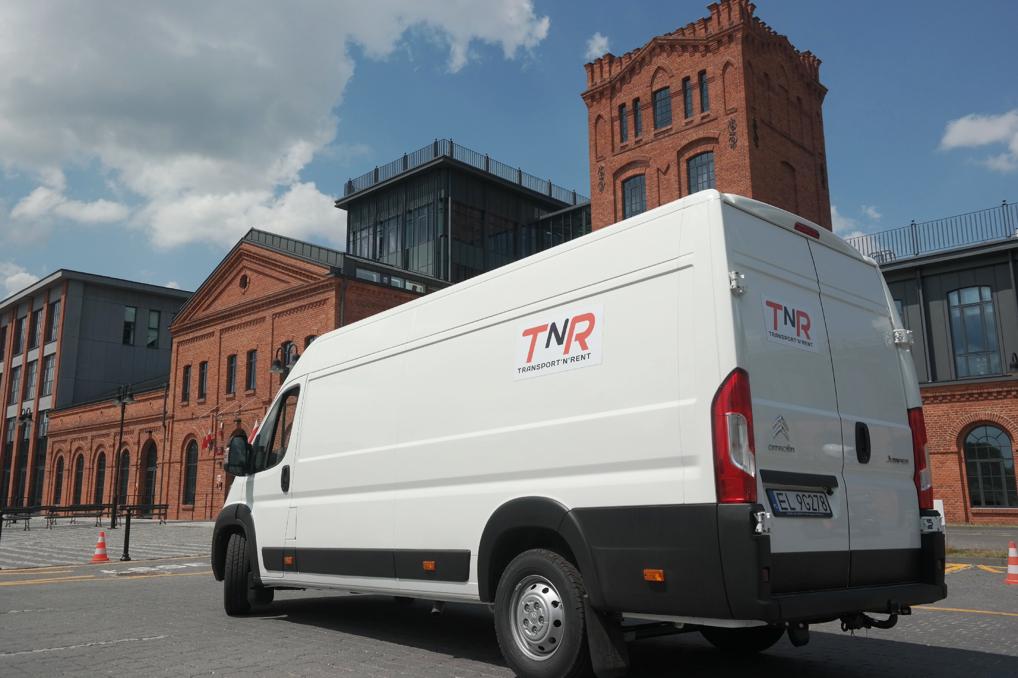 Biały bus z logo firmy TNR Transport'N'Rent zaparkowany na tle ceglanych budynków w słoneczny dzień.