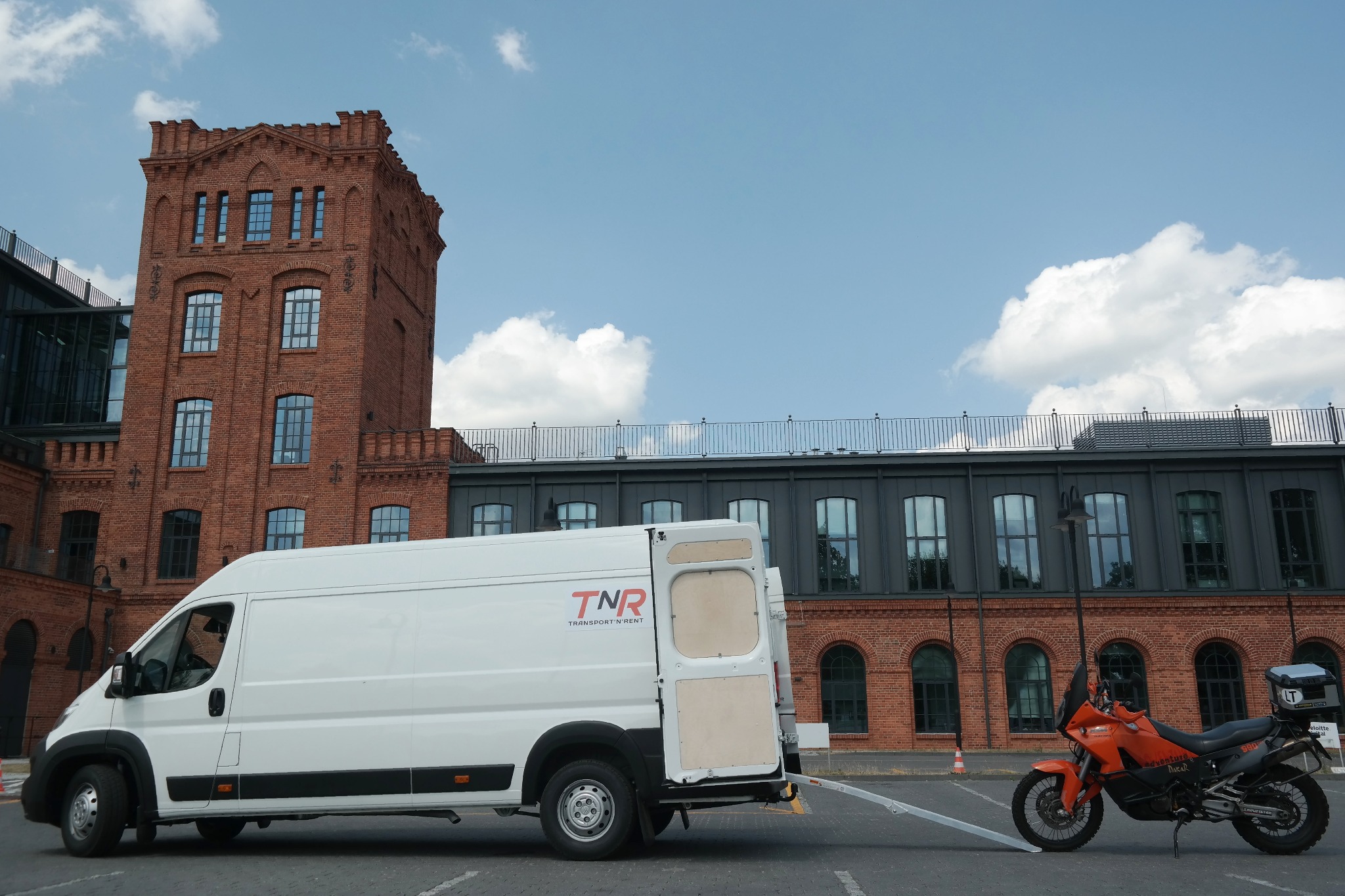 Biały bus z otwartymi tylnymi drzwiami i rampą, obok pomarańczowy motocykl adventure, na tle ceglany budynek