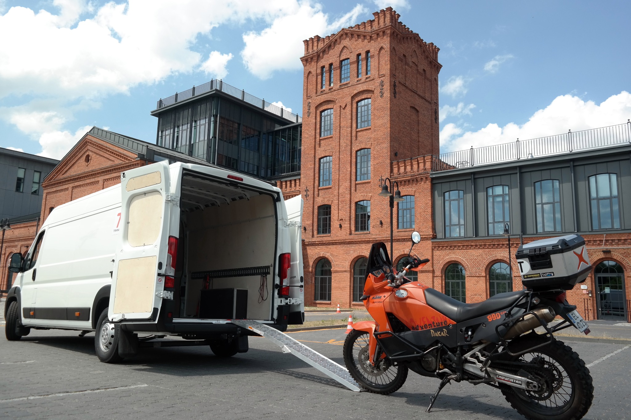 Biały van z otwartymi drzwiami i rampą załadunkową, obok pomarańczowy motocykl KTM Adventure 990 Dakar, w tle budynki z czerwonej cegły.