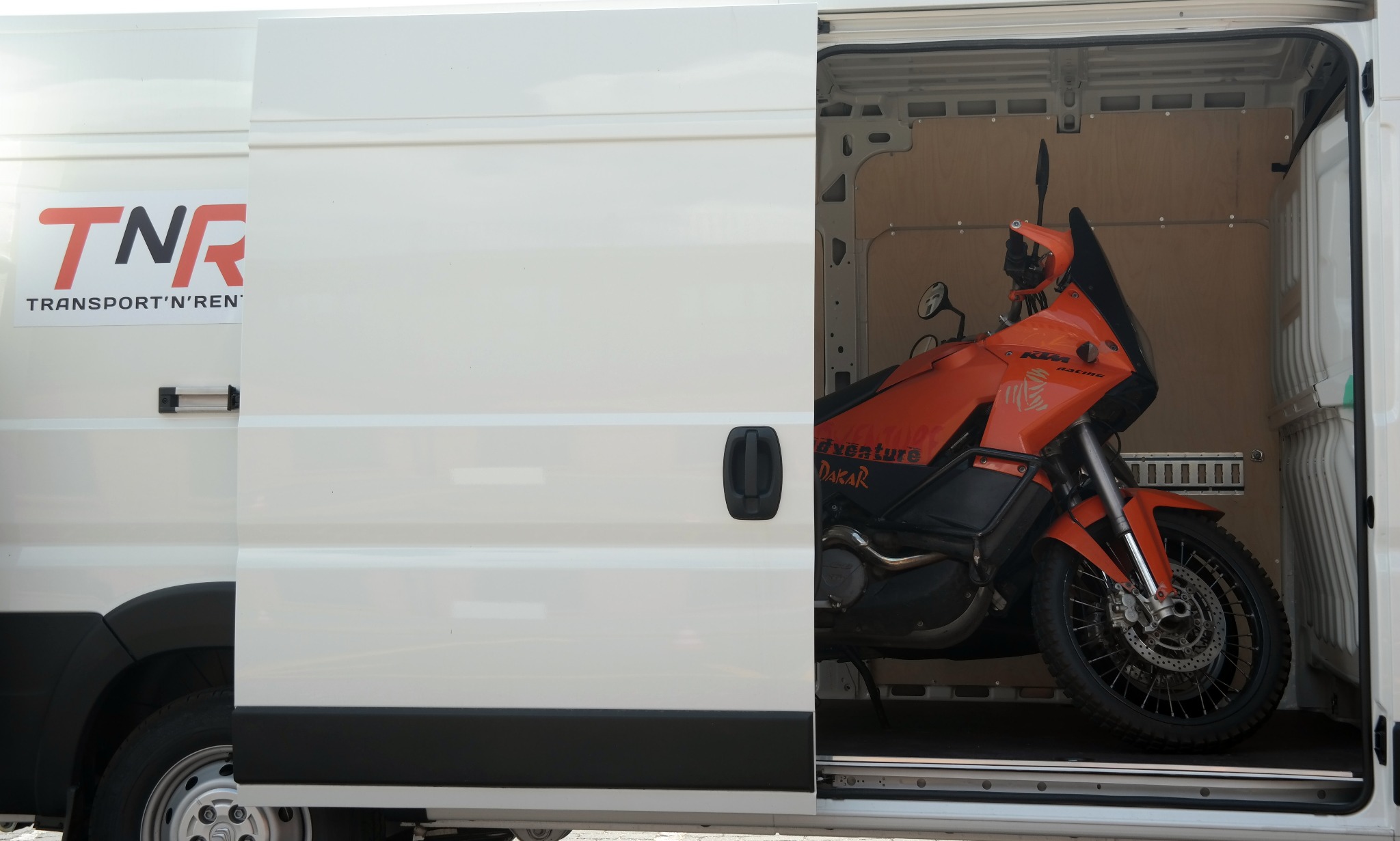 Pomarańczowy motocykl KTM Adventure Dakar zabezpieczony wewnątrz białego busa z częściowo otwartymi drzwiami przesuwnymi, widoczne logo firmy transportowej TNR.