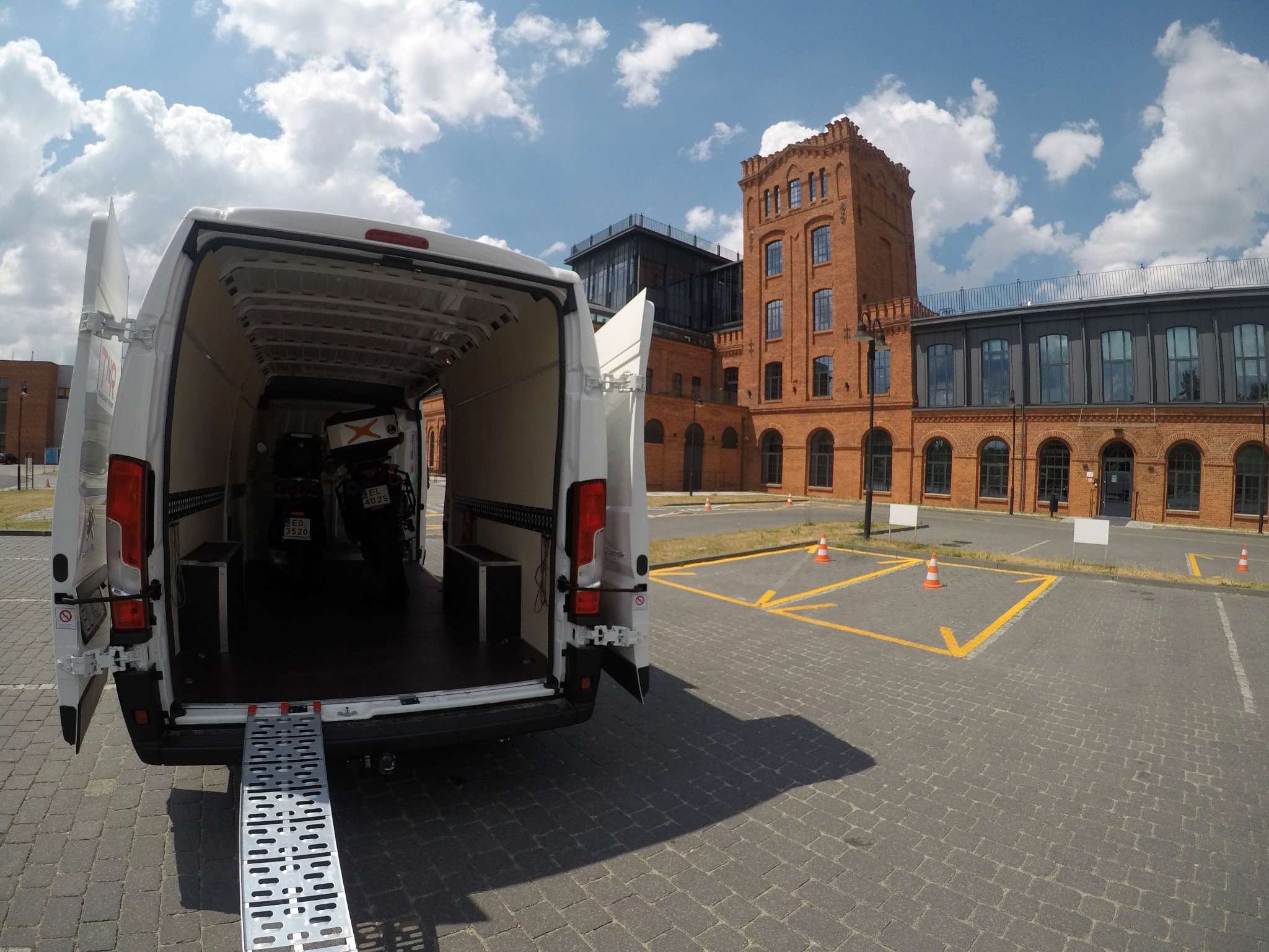 Otwarty biały bus z rampą załadunkową na parkingu, wewnątrz motocykle, w tle ceglany budynek fabryczny.