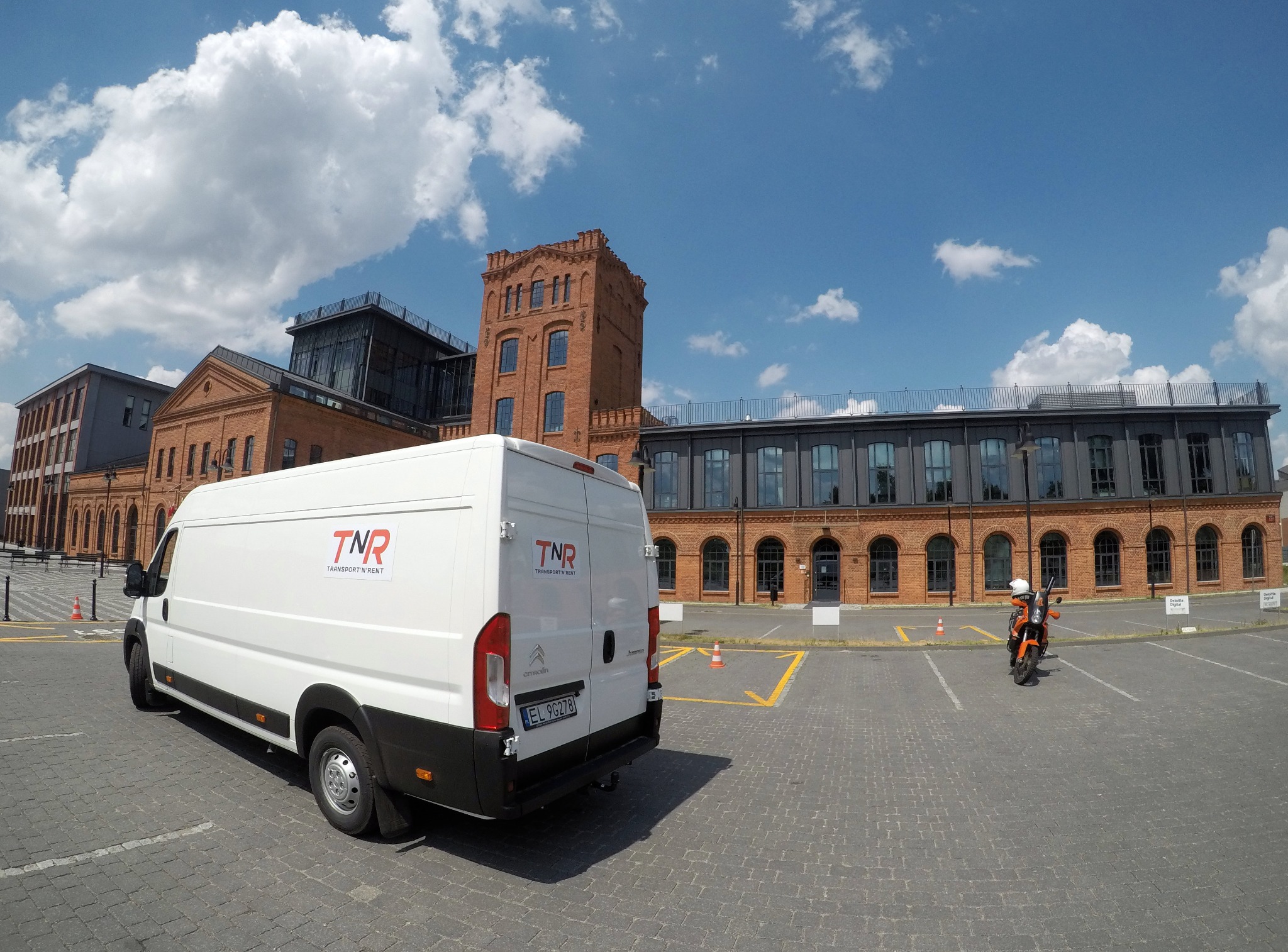 Biały furgon marki Citroen z logo firmy transportowej TNR na tle odrestaurowanych budynków pofabrycznych i motocykla na parkingu.