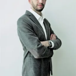 Portret mężczyzny w szarej marynarce z łatami na łokciach, białej koszuli i brązowych spodniach, stojącego na tle jasnej ściany z założonymi rękami.