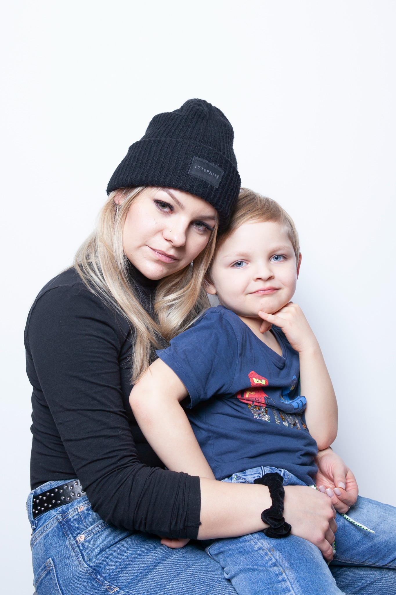Portret matki w czarnej czapce z synem na kolanach, oboje patrzą w obiektyw na białym tle.