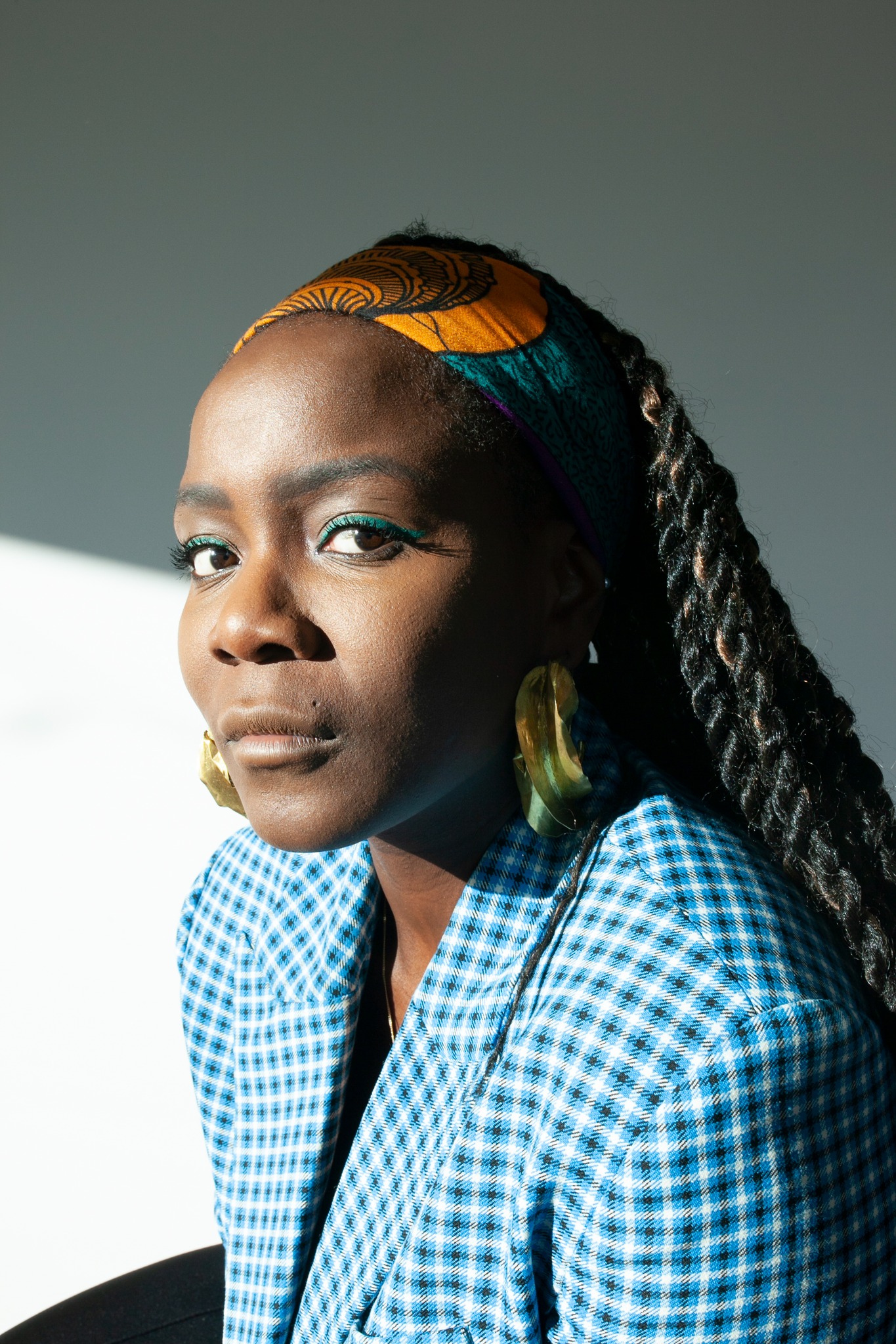 Portret kobiety z warkoczami, w niebieskiej koszuli w kratę, z żółtymi kolczykami i w kolorowej opasce, z zielonym eyelinerem na tle białej ściany.