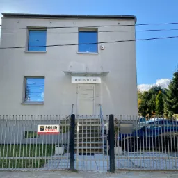 Dwupiętrowy budynek biurowy w Bielsku-Białej z szarą fasadą, oknami odbijającymi niebo i tabliczką 'Biuro Rachunkowe' nad wejściem, ogrodzony metalowym płotem z reklamą firmy ochroniarskiej.