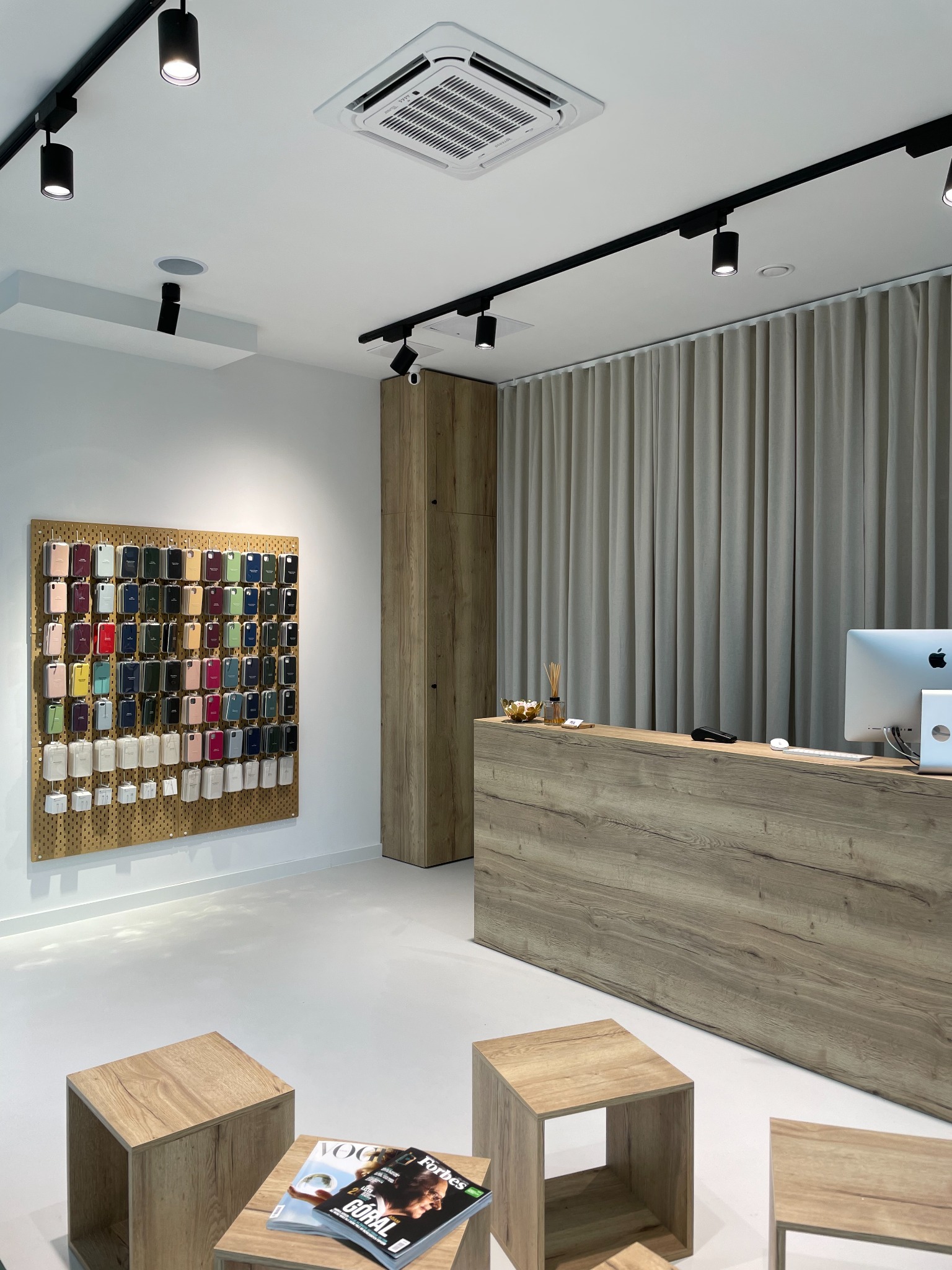 Wnętrze sklepu z akcesoriami do telefonów: drewniana lada z komputerem Apple, ściana z ekspozycją etui na telefony w różnych kolorach, drewniane stoliki z magazynami, oświetlenie punktowe na szynach.