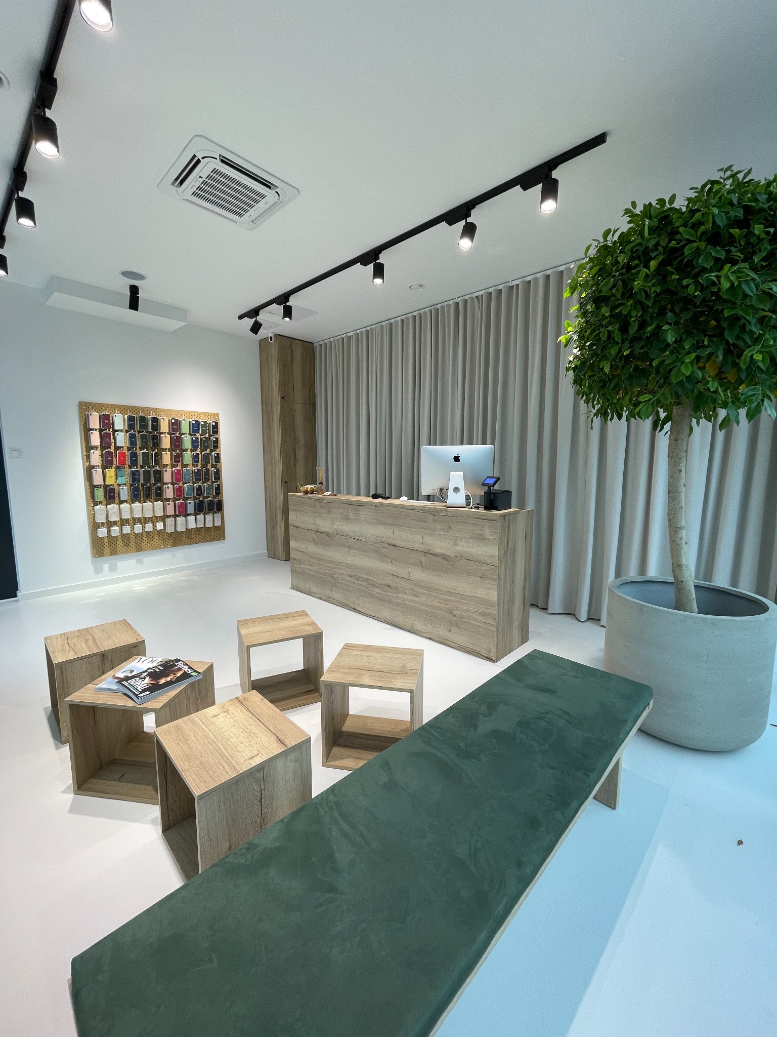 Jasne wnętrze sklepu z akcesoriami GSM, widoczna ściana z ekspozycją etui na telefony, drewniane stoliki i zielona ławka dla klientów, stanowisko obsługi klienta z komputerem iMac.