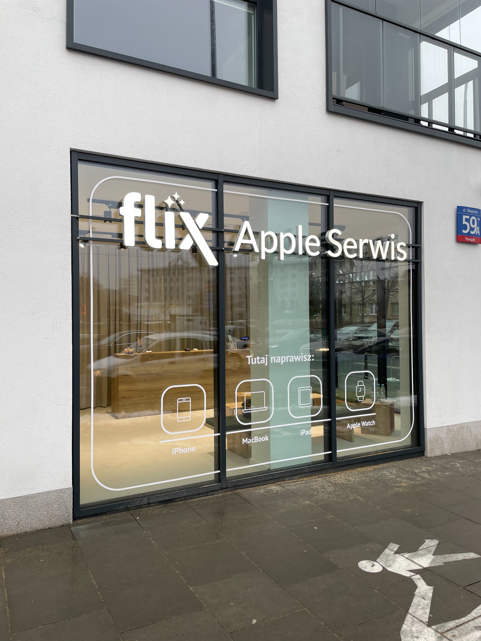 Witryna punktu serwisowego Apple z logo 'flix' i informacją o naprawie iPhone, MacBook, iPad i Apple Watch, widoczna z ulicy Okopowej 59A na Powązkach.