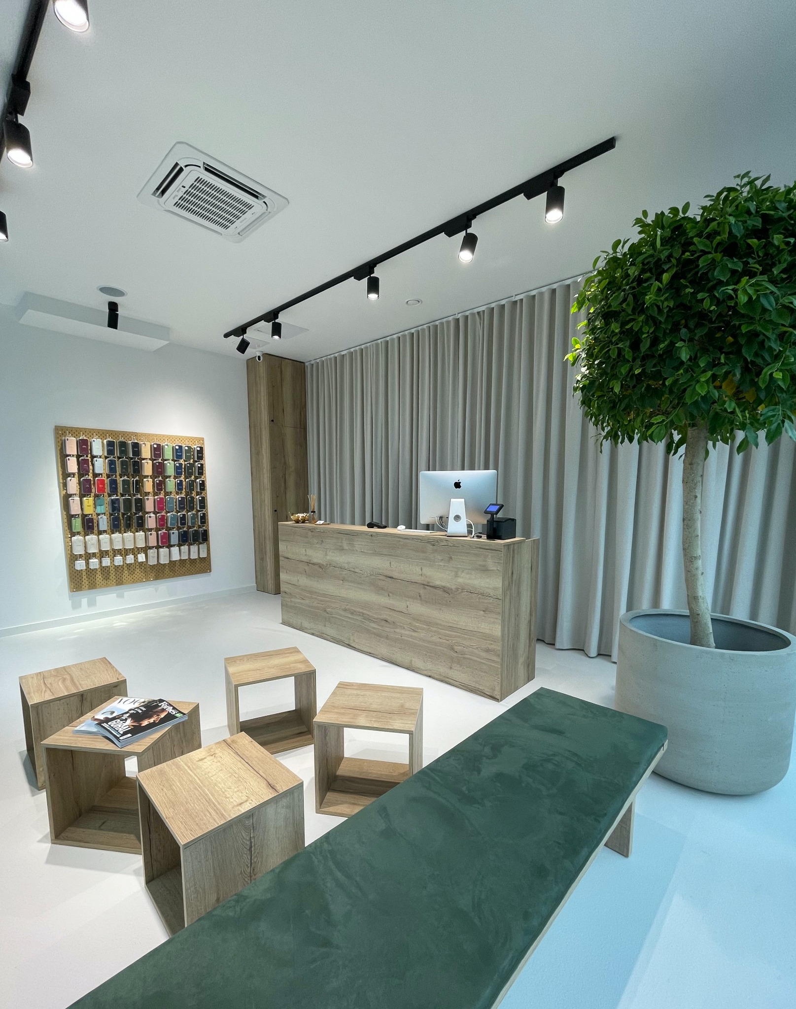 Jasne wnętrze sklepu z akcesoriami do telefonów: drewniana lada z komputerem, ekspozycja etui na ścianie, minimalistyczne meble i roślina w donicy.