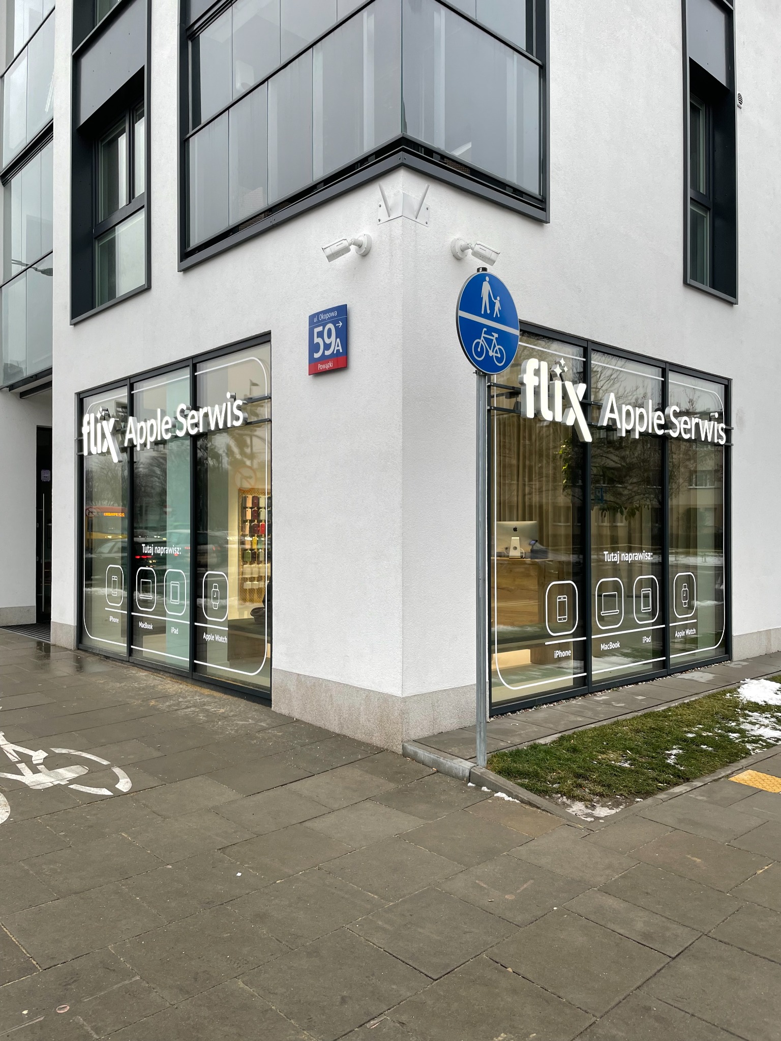 Zewnętrzny widok narożnego budynku z dużymi oknami, na których widnieje napis 'flix Apple Serwis' oraz ikony iPhone, Macbook, iPad i Apple Watch, znak drogowy z symbolem pieszych i roweru...