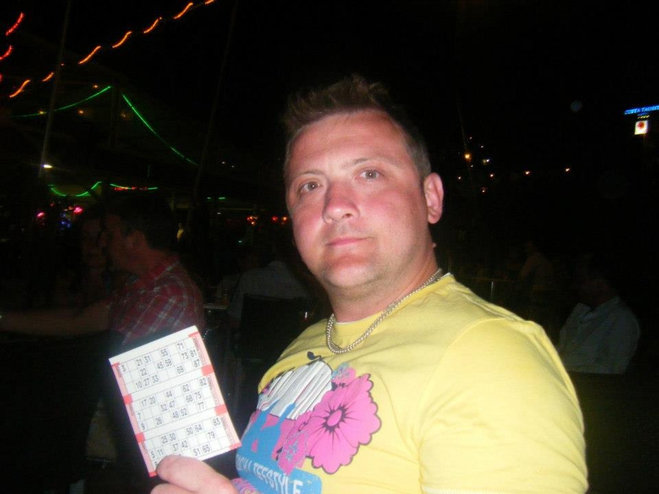 Mężczyzna w żółtej koszulce z kartą do bingo w ręku, na tle ciemnego wnętrza z dekoracyjnym oświetleniem.