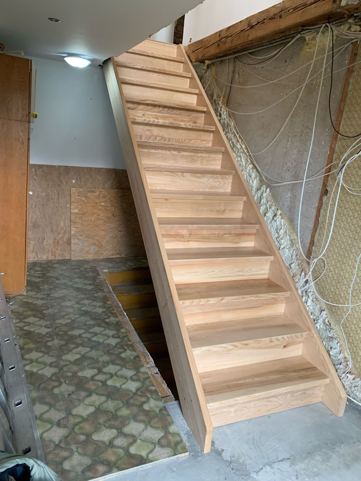 Surowe drewniane schody w trakcie montażu w pomieszczeniu z odsłoniętą ścianą i widocznymi kablami, prowadzące na poddasze.