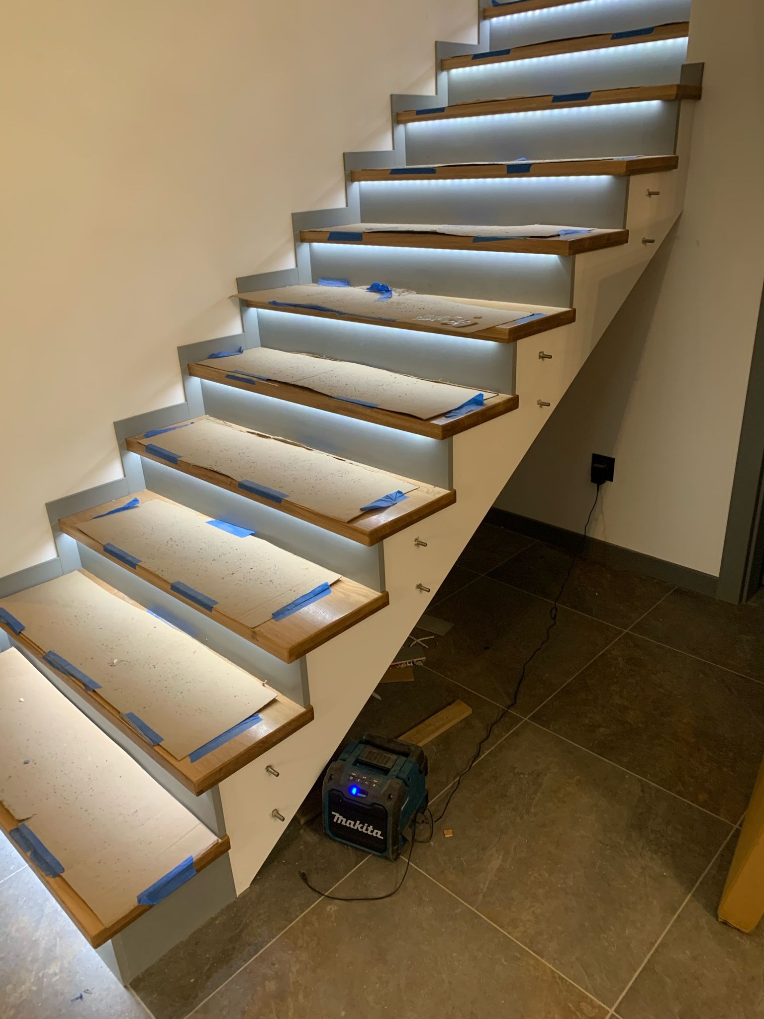 Drewniane schody z podświetleniem LED w trakcie montażu, zabezpieczone kartonem i taśmą malarską, widoczny głośnik budowlany Makita.