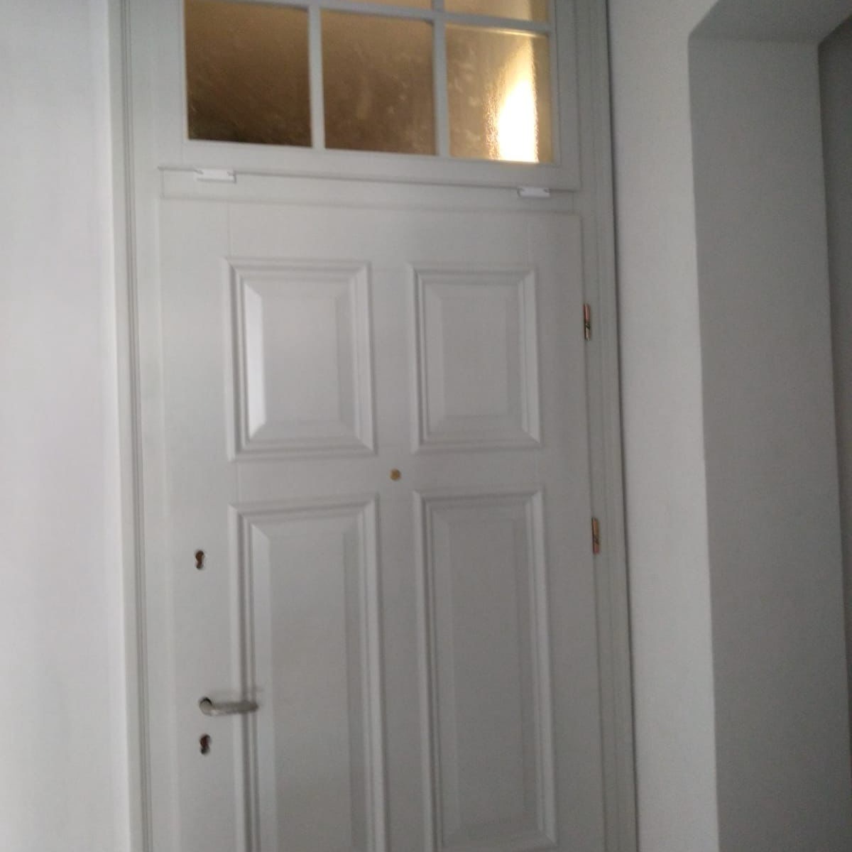 Białe, wewnętrzne drzwi z szybą u góry, widoczne w korytarzu, z metalową klamką i zamkiem.