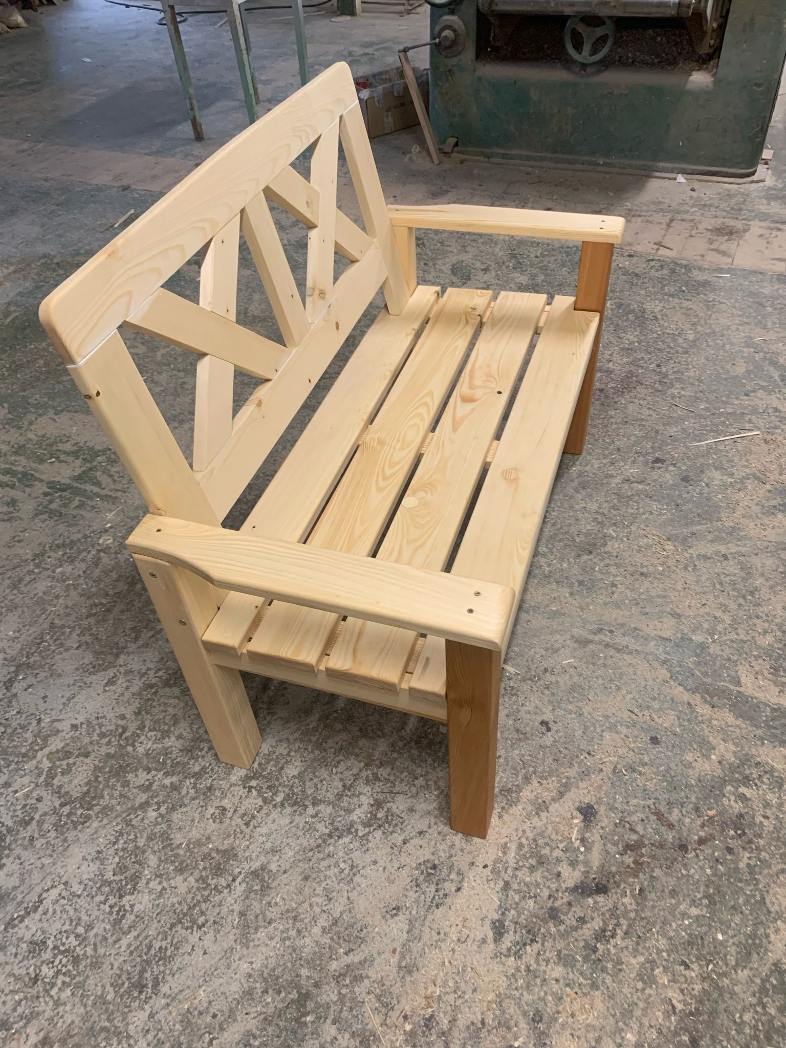 Surowa, drewniana ławka ogrodowa z oparciem w kształcie litery X, wykonana z jasnego drewna, stojąca na betonowej posadzce w warsztacie, z widoczną maszyną stolarską w tle.
