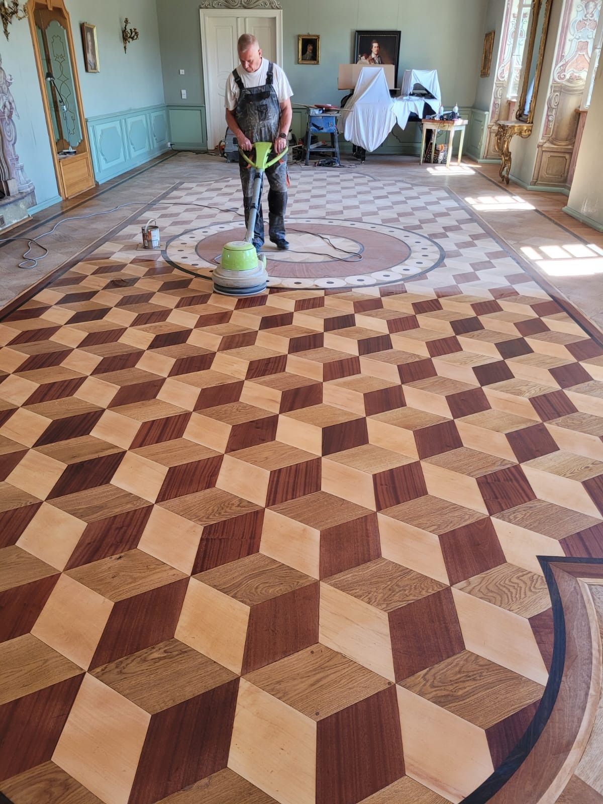 Pracownik w kombinezonie poleruje ozdobny parkiet z efektem 3D w przestronnym, klasycznym wnętrzu pałacowym, używając maszyny do polerowania podłóg.