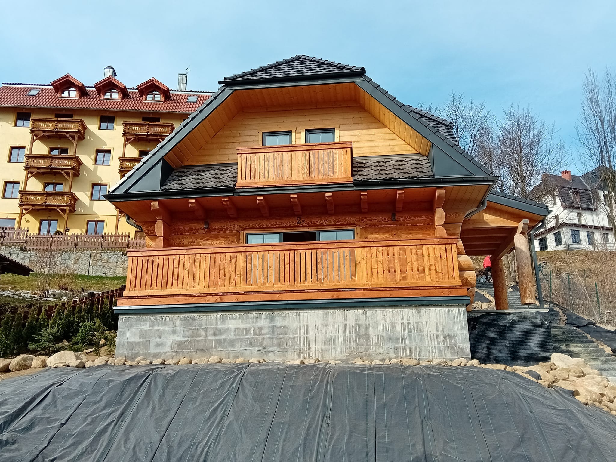 Nowo wybudowany, dwukondygnacyjny dom z bali z drewnianymi balkonami i ciemną dachówką, posadowiony na betonowym fundamencie, otoczony czarną folią budowlaną i kamieniami.