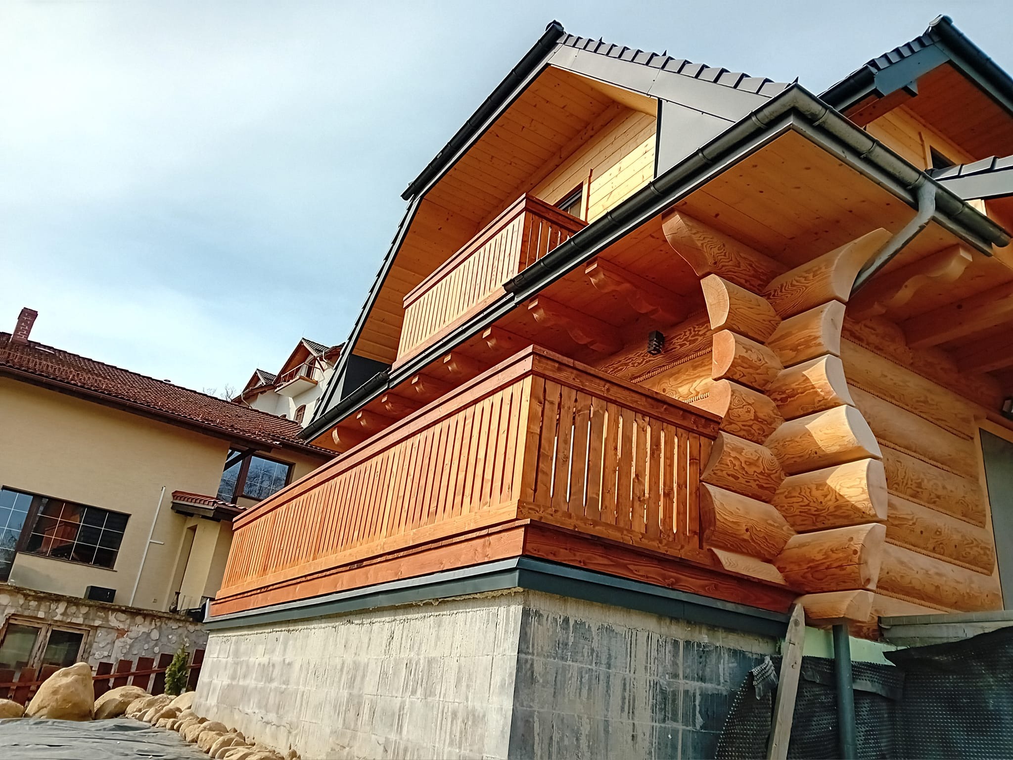 Dom z bali z drewnianym balkonem, widok z dołu, surowy betonowy fundament, w tle inny budynek.