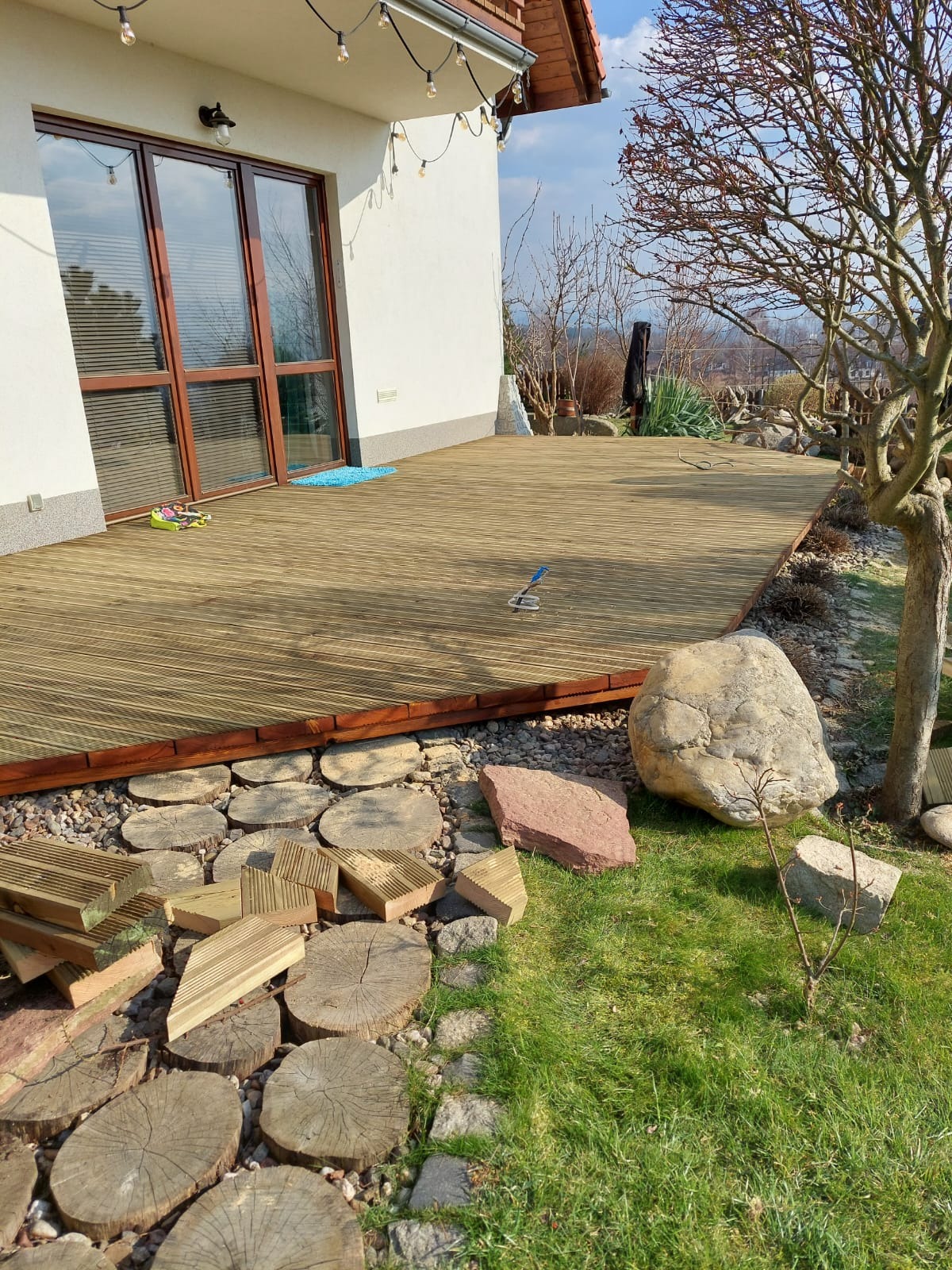 Nowo wybudowany drewniany taras przy domu z elementami dekoracyjnymi z drewna i kamienia w ogrodzie, narzędzie do montażu leży na desce