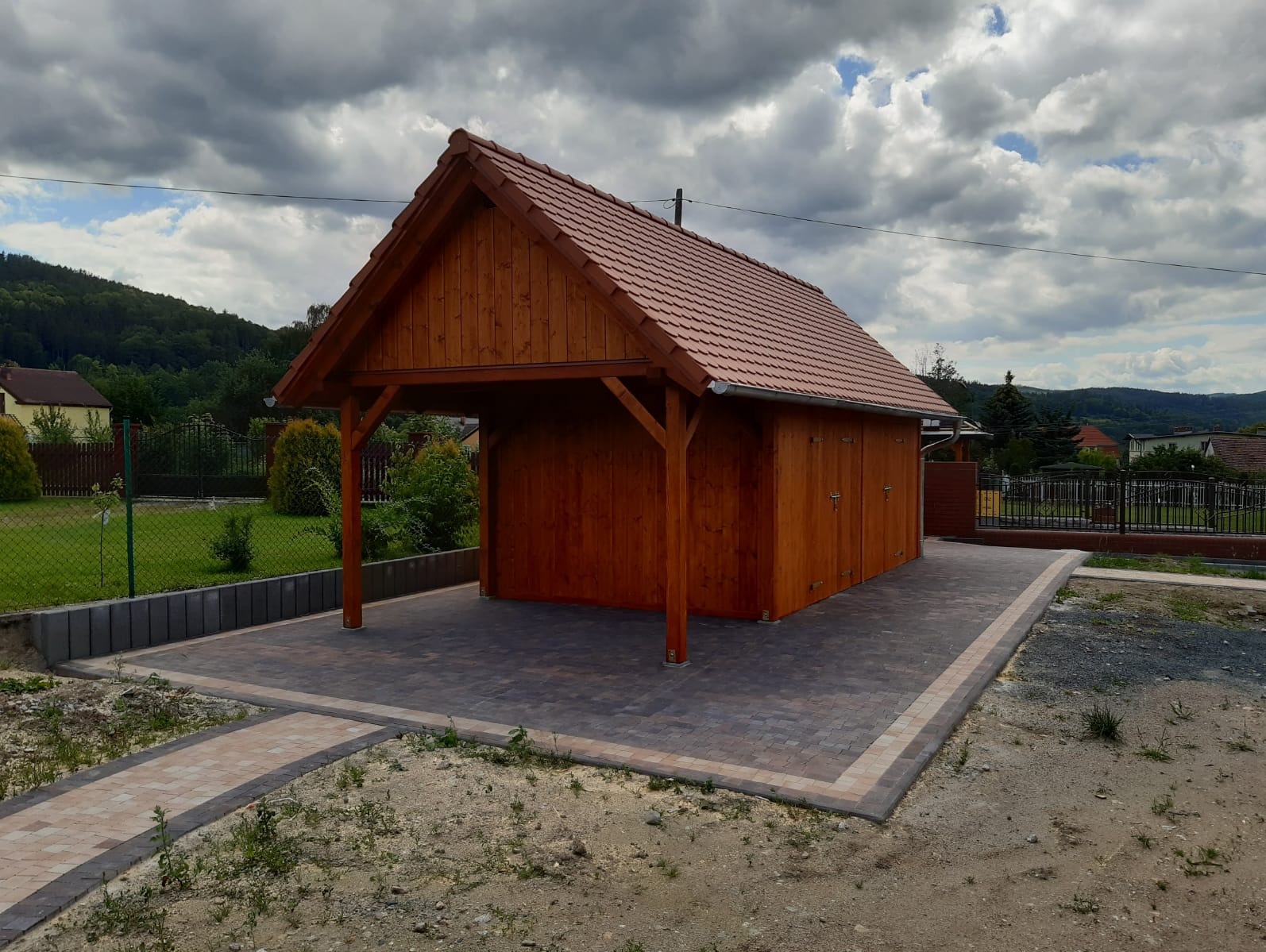 Drewniana konstrukcja z zadaszeniem i zamykanymi drzwiami, wykończona czerwoną dachówką, posadowiona na wybrukowanym placu z kostki w odcieniach szarości i beżu, w tle zielone wzgórza i pochmurne...