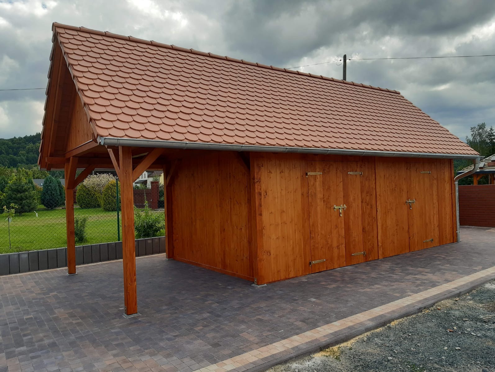 Drewniana konstrukcja z dwoma parami drzwi, kryta dachówką ceramiczną, z otwartą wiatą z boku i brukowanym podjazdem.