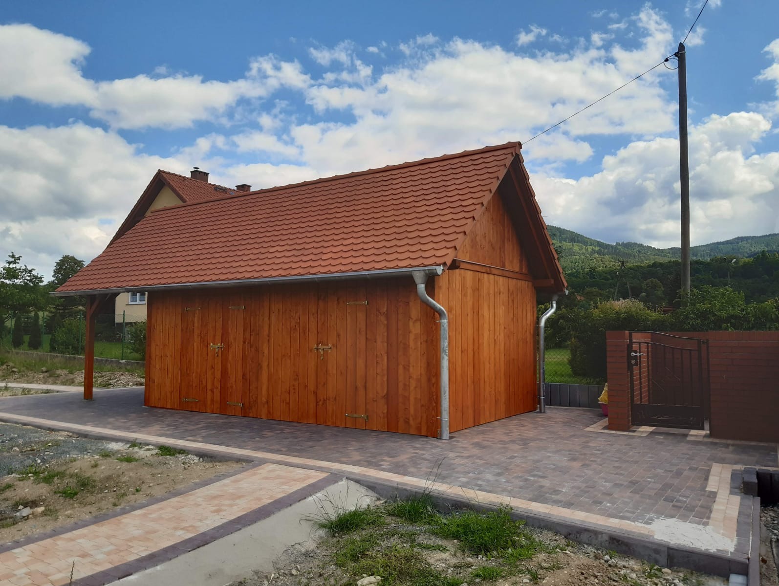 Drewniany garaż z czerwonym dachem, widoczne metalowe rynny, brama wjazdowa i częściowo ułożona kostka brukowa na podjeździe, w tle zielone wzgórza i błękitne niebo z chmurami.