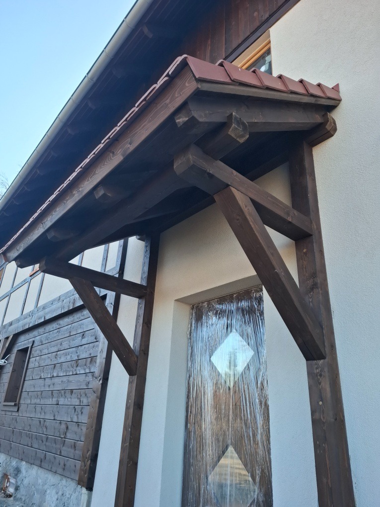 Drewniana wiata nad wejściem do domu z krytą konstrukcją wspartą na słupach, drzwi zabezpieczone folią ochronną.