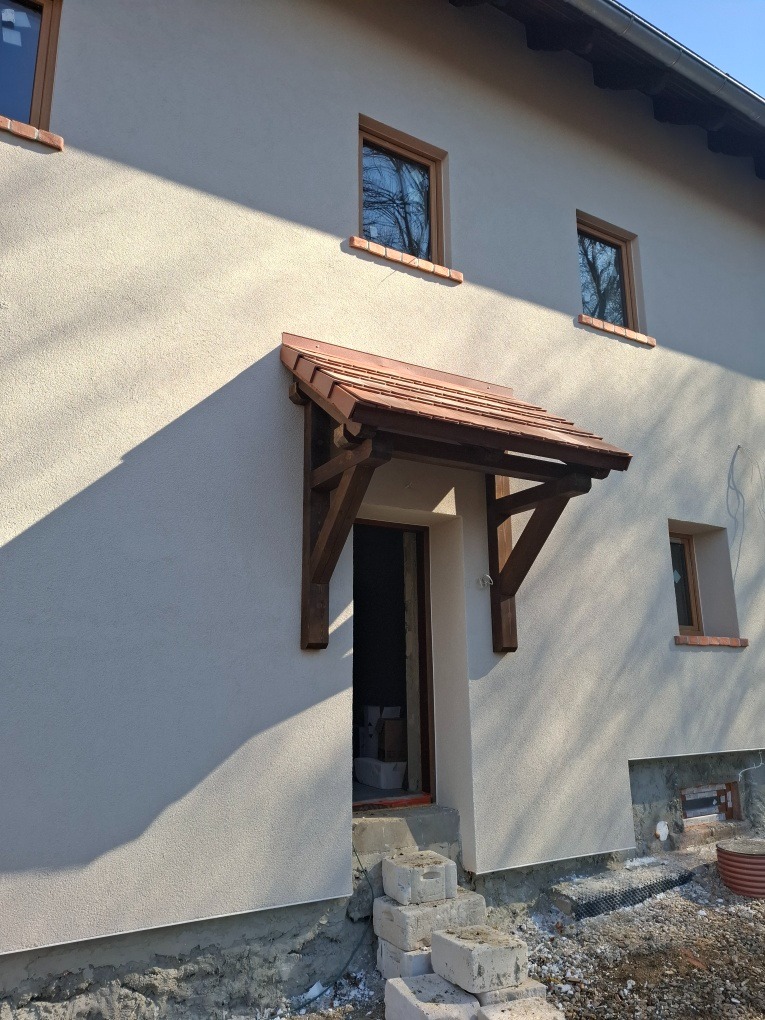 Elewacja domu z nowym oknem i drewnianym zadaszeniem nad wejściem, widoczne tymczasowe schody z bloczków betonowych.